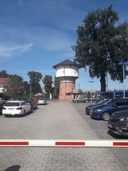teilweise ungenutzte Stehblechplätze hinter einer Schranke; der Boden ist vollversiegelt. lm Hintergrund ein einzelner großer Baum und ein alter Wasserturm.